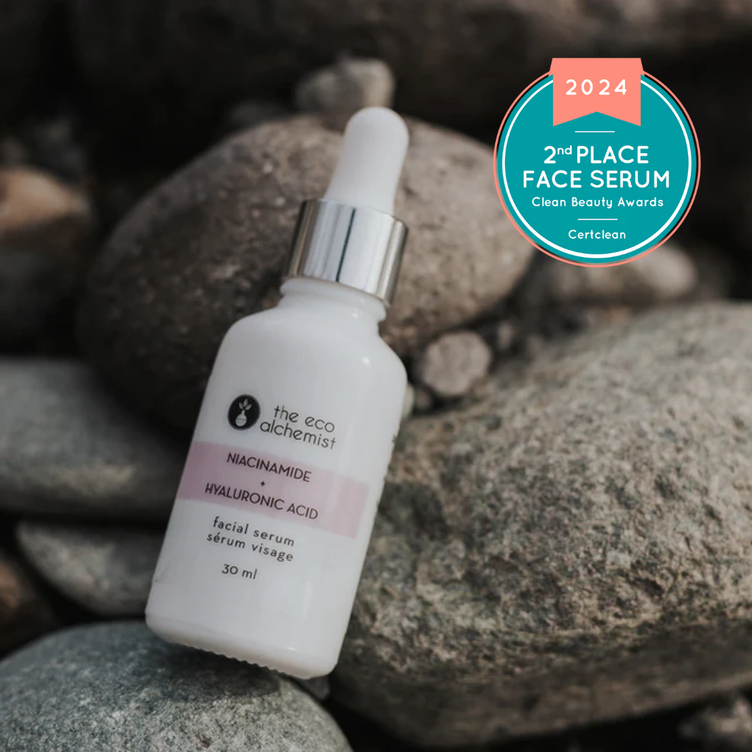 A bottle of Niacinamide + Hyaluronic Acid Hydrating Facial Serum by The Eco Alchemist, placed against a backdrop of rocks, with a 2024 second place face serum award badge displayed.