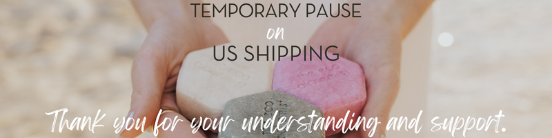 A close-up of two hands holding three shampoo bars in beige, green, and pink, with overlaid text reading 'TEMPORARY PAUSE ON US SHIPPING' and 'Thank you for your understanding and support.'.